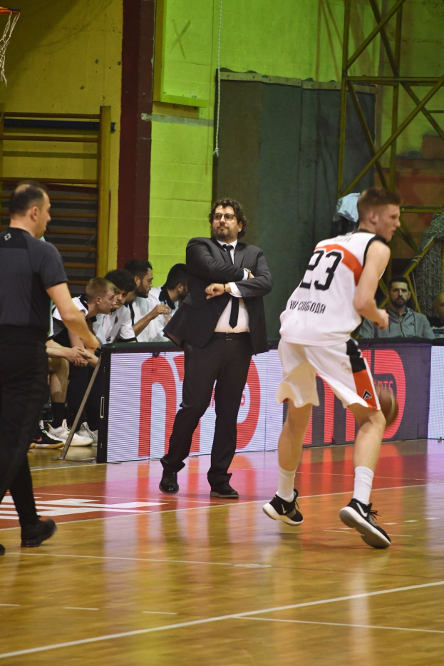 Eine Gruppe von Männern spielt Basketball auf einem Court mit Zuschauern im Hintergrund und einem Basketballkorb in der oberen linken Ecke, alle tragen Sneaker.