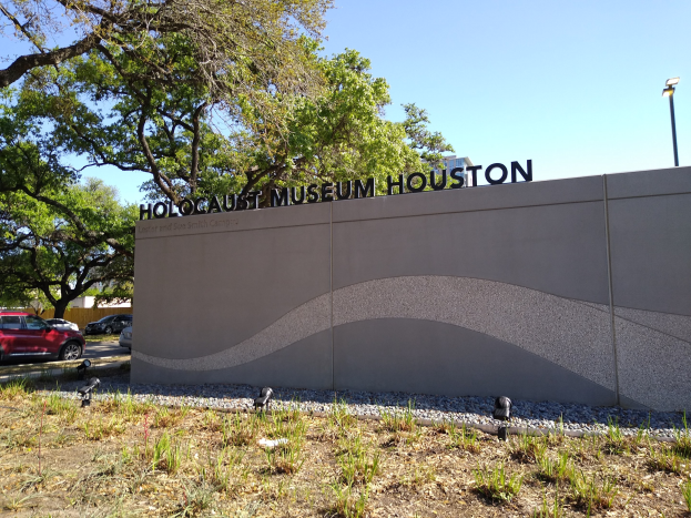 Außensicht des Holocaust-Museums in Houston mit einer Gedenkwand, Steinen, Pflanzen, Straßenfahrzeugen, einem Straßenpfahl und einem bewölkten Himmel.