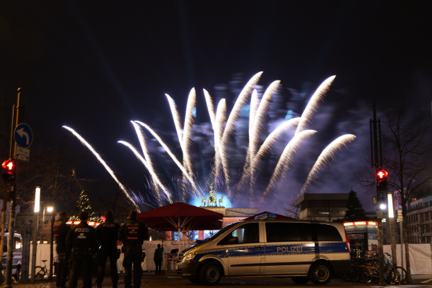 Polizeifahrzeug vor einer Menge mit farbigen Feuerwerken am Himmel und beleuchteten Gebäuden.