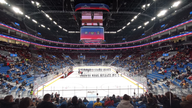 Große Halle mit Zuschauern bei einem Hockey-Spiel, mit einer Gruppe von Menschen in einem eingezäunten Bereich, Schildern, Metallrahmen, einem Display und Deckenleuchten.