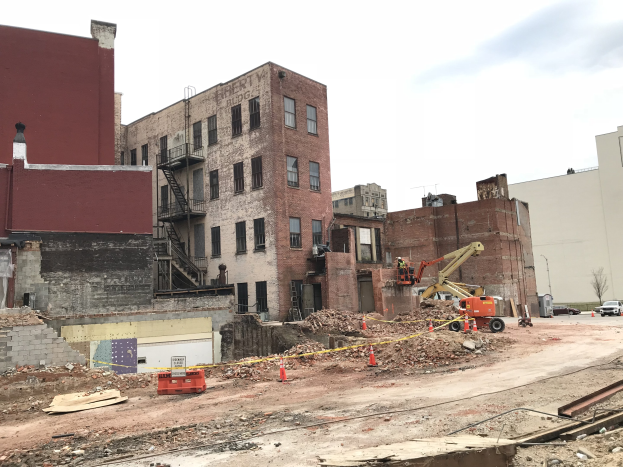 Baustelle mit einem großen Gebäude im Hintergrund, umgeben von Fahrzeugen, Verkehrskegeln, Pfählen und Bäumen an einem bewölkten Tag.