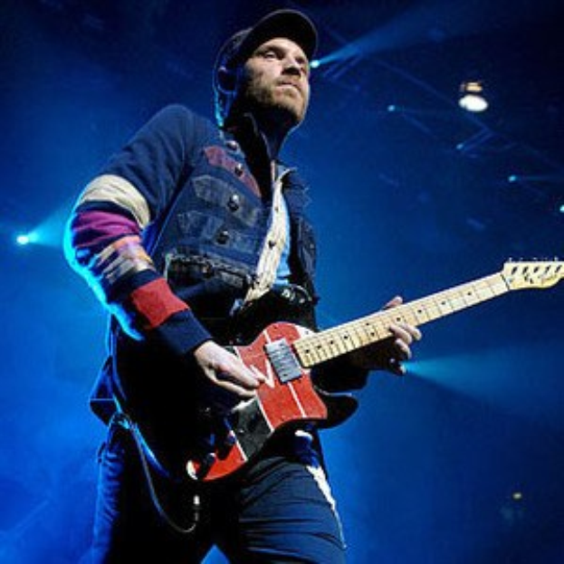 Ein Mann mit einer Mütze spielt eine E-Gitarre auf einer hell erleuchteten Bühne und strahlt vor Freude und Aufregung.