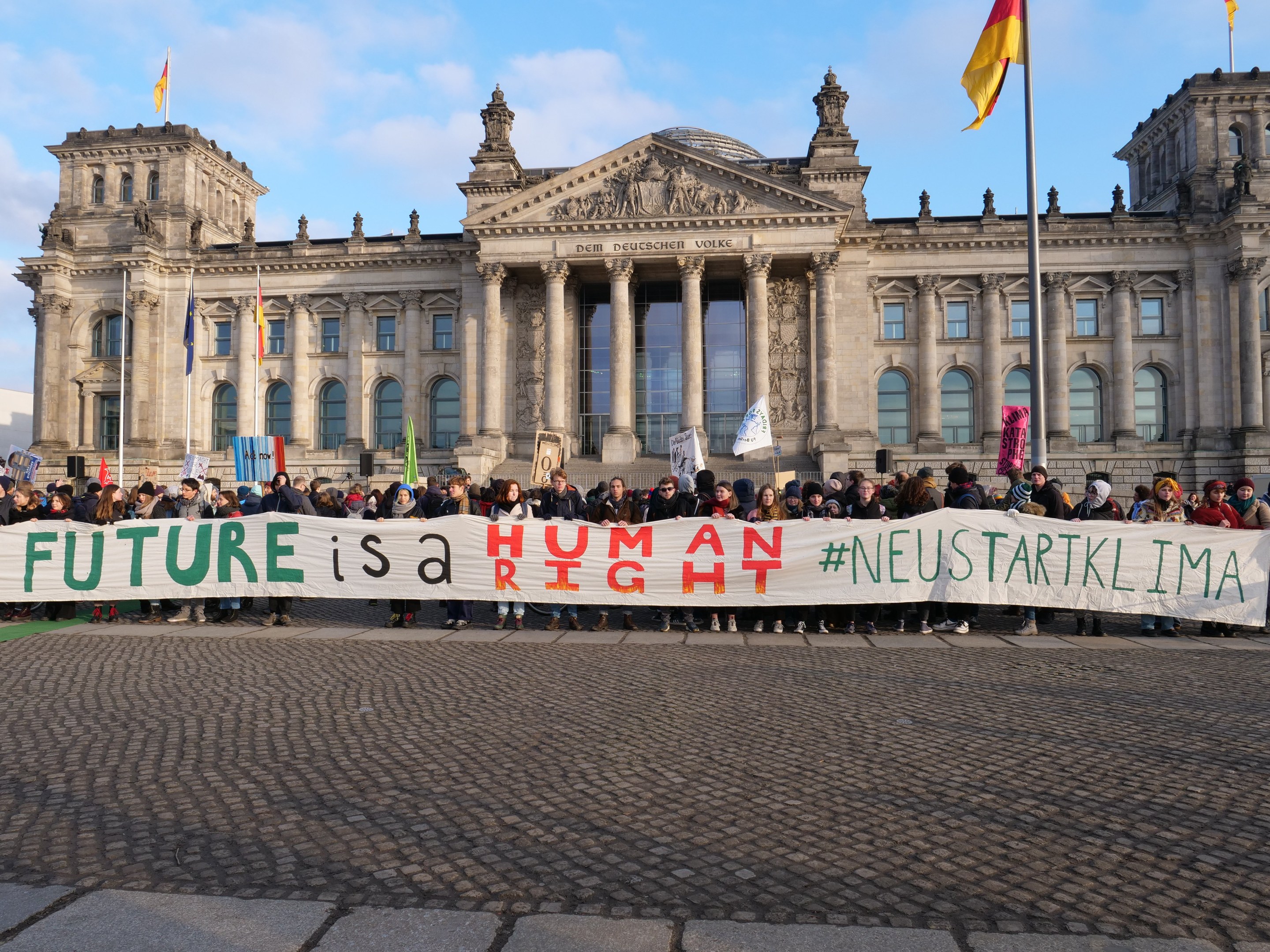 Eine Gruppe von Menschen hält ein Transparent mit der Aufschrift 'Zukunft ist ein Mensch' vor dem Reichstaggebäude in Berlin, Deutschland, mit sichtbaren Fenstern, Säulen und Bögen des Gebäudes, umgeben von Fahnenmasten und einem bewölkten Himmel.