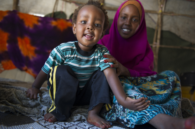 Eine Frau und ein Kind sitzen unter einem drapierten Tuch in einem Zelt, mit Holzstöcken und einer Wand im Hintergrund; die Frau hat ausgestreckte Arme und einen entschlossenen Gesichtsausdruck.