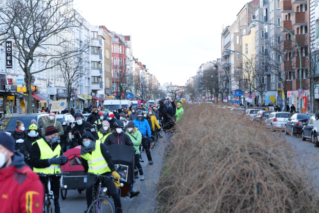 Eine große Gruppe von Menschen in Masken und Sicherheitswesten auf Fahrrädern auf einer von Bäumen gesäumten Straße mit Gebäuden, Fahrzeugen, Laternen und Texttafeln, vor einem klaren blauen Himmel.