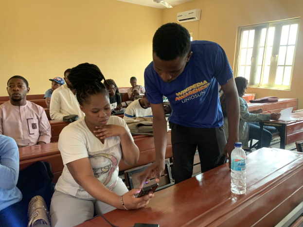 Eine Gruppe von Schülern sitzt an Tischen in einem Klassenzimmer, während ein Mann mit einem Mobiltelefon in der Hand steht; Flaschen und andere Gegenstände sind auf den Tischen, und eine Klimaanlage und ein Fenster sind im Hintergrund zu sehen.