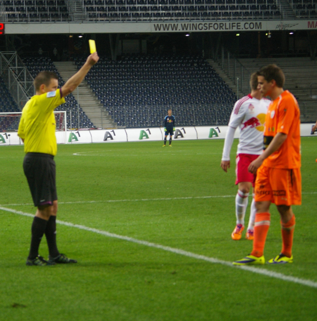 Ein Schiedsrichter hält eine Gelbe Karte in Richtung eines Fußballspielers, während andere in der Nähe auf dem Feld stehen, mit einem Torpfosten, einem Zaun, einem Schild, Metallrahmen, einem Anzeigebildschirm, Stühlen und Deckenlampen im Hintergrund.