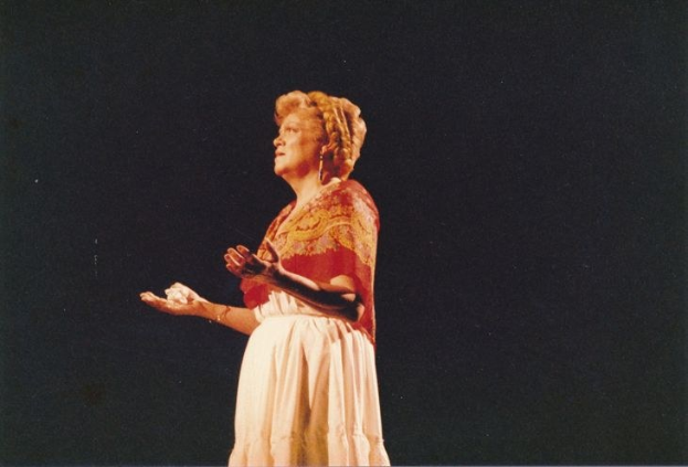 Eine Frau in einem langen weißen Kleid mit hohem Kragen und langen Ärmeln steht auf einer Bühne mit ausgestreckten Armen, ihr Haar ist zurückgebunden und sie hat einen ernsten Gesichtsausdruck, vor einem dunklen Hintergrund.