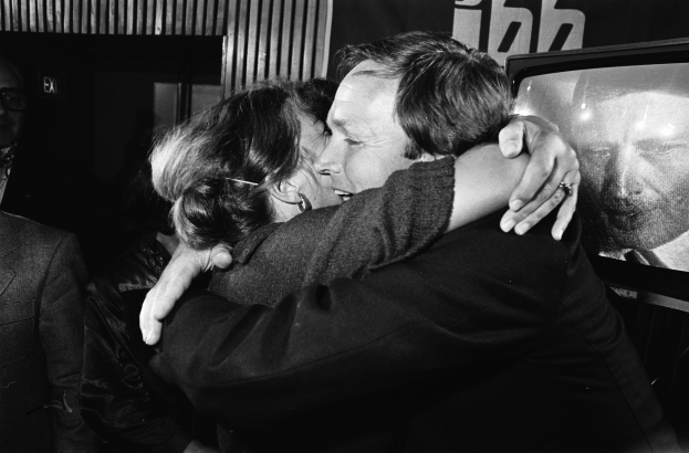 Black and white image of a man in a suit and a woman in glasses embracing in front of a television, with a banner in the background.