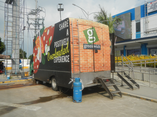 Lastwagen vor einem Gebäude mit einer Tafel, auf der "Teilen Sie eine positiv überladene Erfahrung" steht, mit Stufen, einem Zylinder auf dem Boden, Geländern, Strommasten mit Drähten, Laternenpfählen, Bäumen und einer bewölkten Wolkenformation.