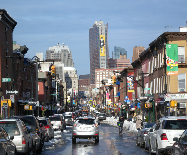 Eine belebte Stadtstraße mit Fahrzeugen, Fahrrädern, Fußgängern, schneebedecktem Boden, Gebäuden, Laternen, Verkehrsampeln, Verkehrsschildern, Bannern und einem bewölkten Himmel.