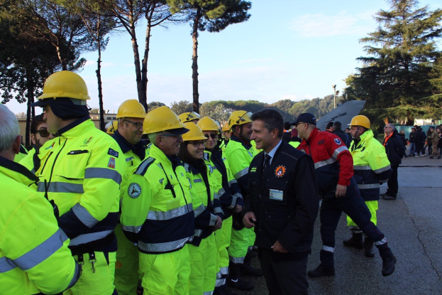 Eine Gruppe von Einsatzkräften in gelben Westen und Helmen, die auf einer Straße mit Bäumen und einem klaren blauen Himmel im Hintergrund stehen.