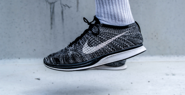 Schwarze und weiße Nike Flyknit Racer-Schuhe mit weißer Zwischensohle und Socken vor einer Wand.