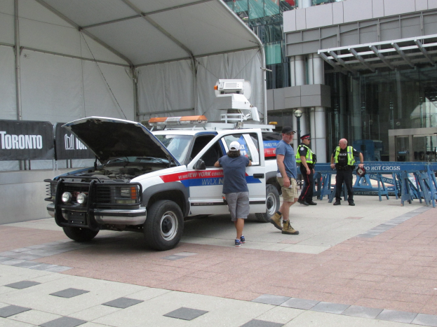 Polizeiauto mit offener Motorhaube vor einem Gebäude geparkt, umgeben von mehreren Personen und Schildern auf der linken Seite.