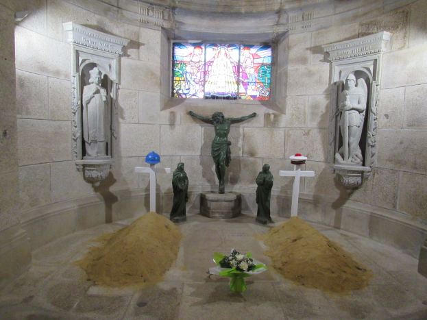 Ein Krypta mit einer zentralen Statue des Jesus am Kreuz, umgeben von Sand und einem Blumenbouquet, mit skulptierten Wänden und einem Buntglasfenster.