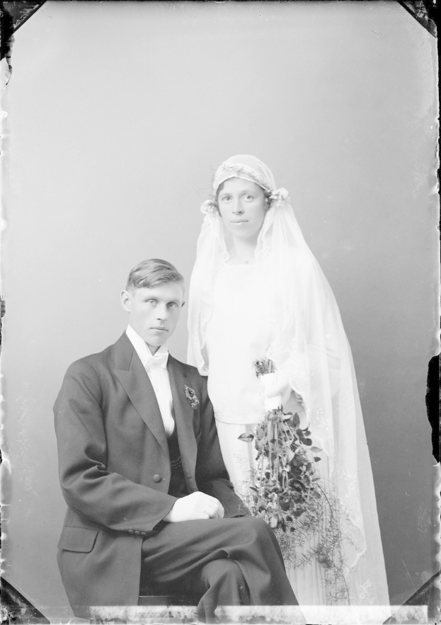 Schwarz-weiß-Foto einer formell gekleideten Braut in einem weißen Kleid und Schleier, die einen Strauß hält, neben einem ebenfalls formell gekleideten Bräutigam sitzend.