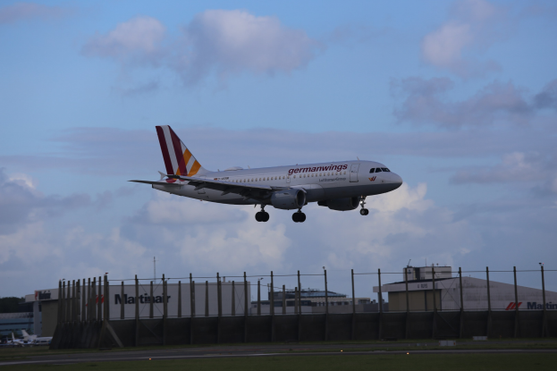 Germanwings Airbus A320-200 beim Abheben vom Frankfurt Airport mit Gras darunter, einem Zaun im Vordergrund, Gebäuden und Bäumen im Hintergrund und einer bewölkten Himmel.