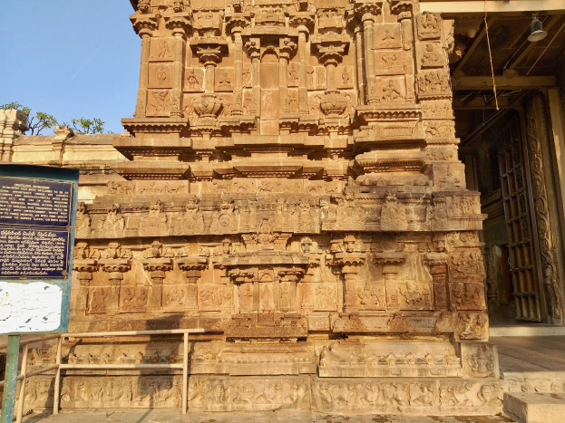 Großer steinerner Khajuraho-Tempel in Indien mit detaillierten Schnitzereien, ein Schild auf einem Ständer, ein Metallzaun, umgebende Bäume und ein bewölkter Himmel.