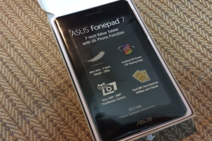 Ein Smartphone mit Spezifikationen auf dem Bildschirm, das in einer weißen Box auf einer braunen Oberfläche liegt.
