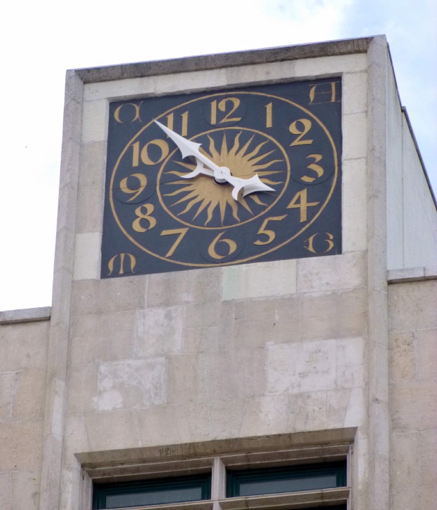 Ein Sonnenuhr-Ziffernblatt, das an der Seite eines Gebäudes mit Fenstern angebracht ist und die Zeit mit zwei entgegengesetzten Händen anzeigt.