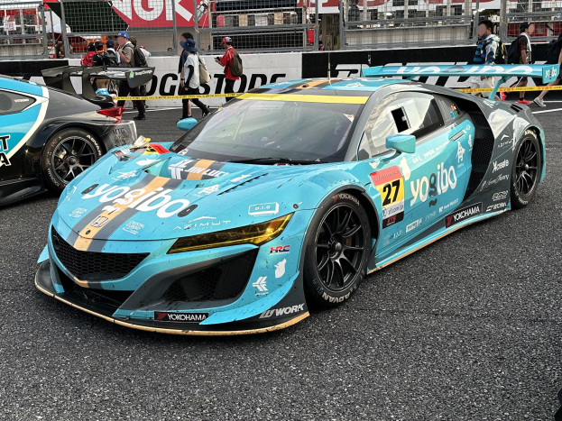 Eine Gruppe von Rennwagen auf einer Rennstrecke mit Menschen in Mützen und Taschen im Hintergrund, umgeben von Texttafeln, Geländern, Pfosten und einem entfernten Gebäude, mit der Beschriftung "Yokohama Acura NSX GT3" unten.