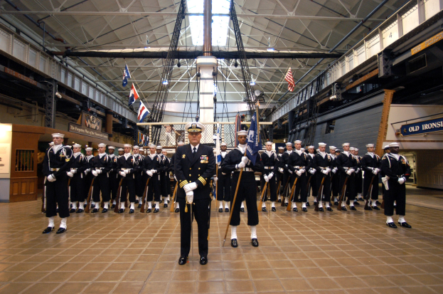 Gruppe uniformierter Männer in einem großen Raum mit Gewehren, die Kappen tragen, mit Fahnen und Schildern an den Wänden, Deckenleuchten und Stangen.