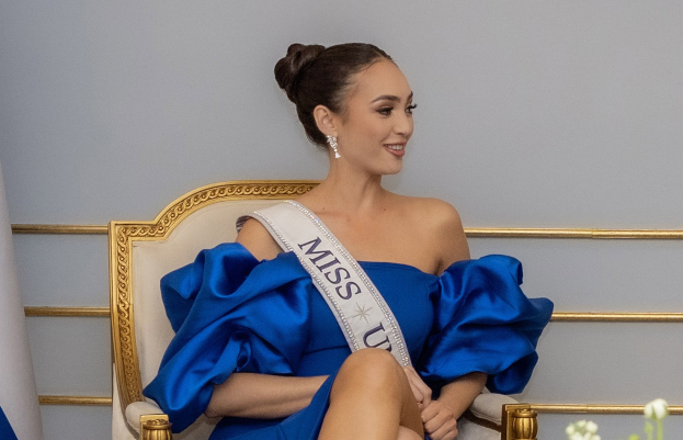 Frau in einem blauen Kleid mit einem "Miss Universe 2019" Band, die auf einem Stuhl sitzt und lächelt.