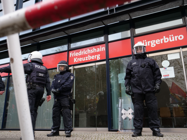Polizisten in Helmen und Handschuhen stehen vor einem Gebäude mit Glaswänden und -türen, mit einem Pfahl links und Schildern mit der Aufschrift "Friedrichshain Kreuzberg" im Hintergrund.