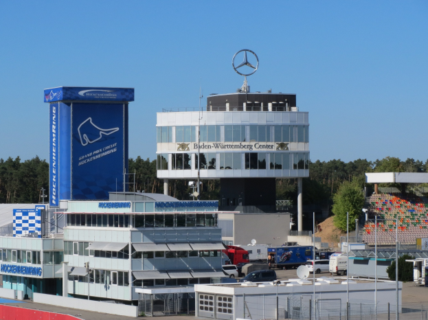 Exterior view of the Mercedes-AMG Petronas F1 Team headquarters in Berlin, Germany, showing buildings, vehicles, a fence, signage, trees, and a logo on the structure, with the sky in the background.