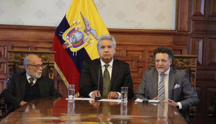 Drei Männer in Blazern und Krawatten sitzen an einem Tisch mit Gläsern, Papieren und Mikrofonen vor einer venezolanischen Flagge und einer hölzernen Wand, scheinbar in einer Besprechung mit José Manuel Barroso, dem Präsidenten von Venezuela, in der Mitte.
