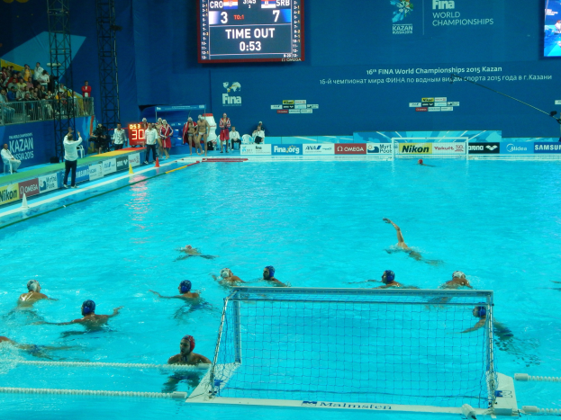 Gruppe von Menschen, die Wasserball in einem Pool während der FINA-Weltmeisterschaften 2015 in Kazan spielen, mit Zuschauern und Veranstaltungsbeschilderung im Hintergrund.