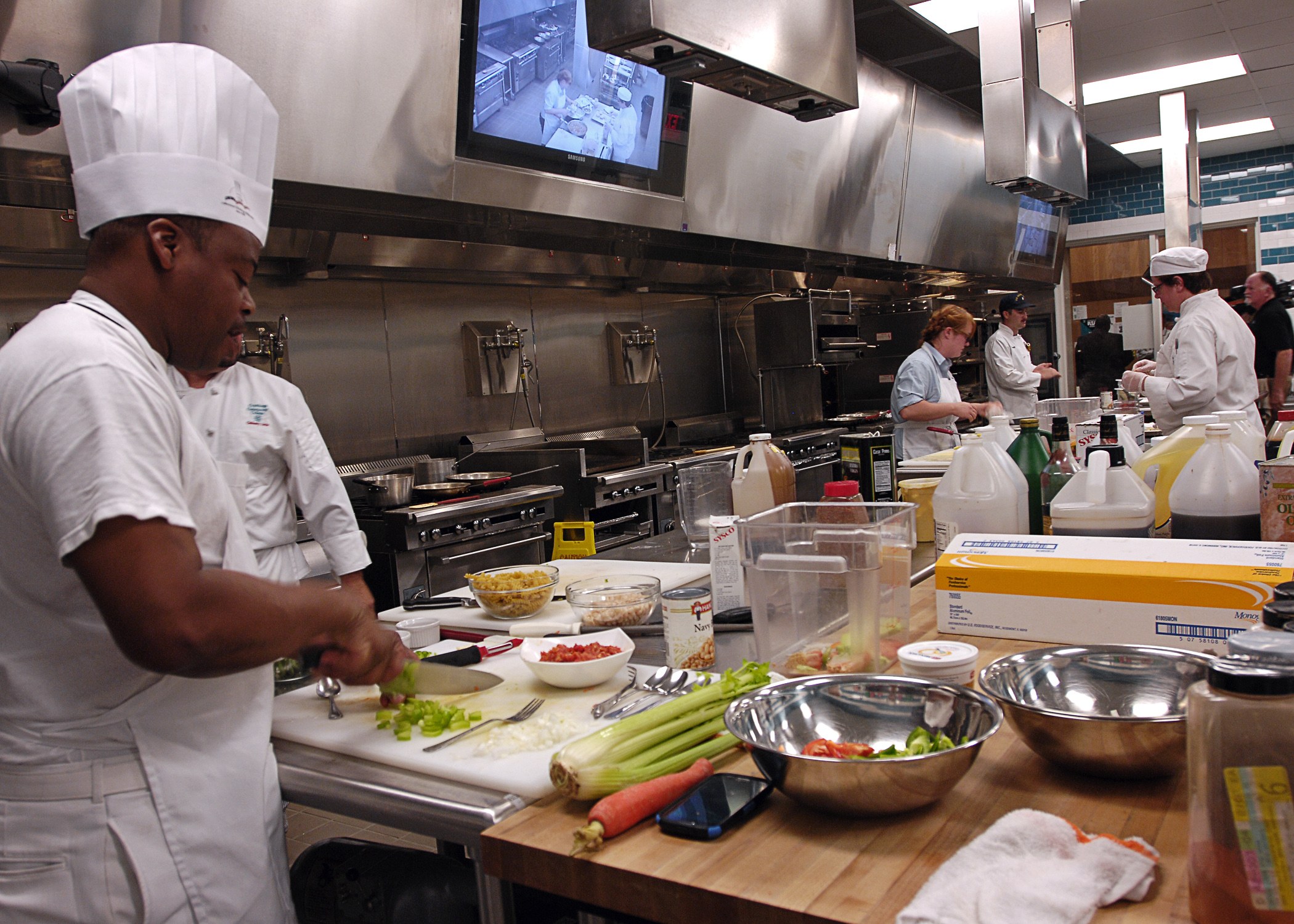 Köche in einer Küche beim Zubereiten von Essen, mit einem in einer weißen Uniform und einer Mütze, der ein Messer hält, umgeben von Utensilien, Gemüse, einem Telefon und Flaschen, mit einem Fernseher und Deckenlampen im Hintergrund.