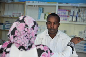 Ein Apotheker in einem weißen Kittel spricht mit einer sitzenden Frau in einer Apotheke, mit Regalen voller Schachteln und anderen Gegenständen dahinter.