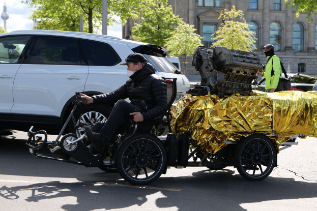 A man in a wheelchair with a large engine on its back, surrounded by vehicles on a road with trees, buildings, and a clear blue sky in the background, wearing a black jacket and cap while holding an object.