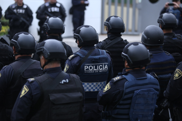 Mexikanische Polizisten in Uniform und Helmen stehen vor einem Gebäude, mit ein paar Menschen und einem Kameramann im Hintergrund, Pflanzen und einem Tor.