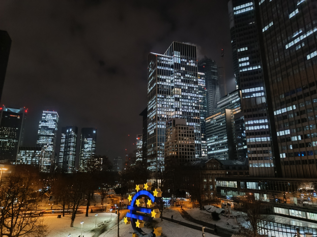 Hauptsitz der Europäischen Zentralbank in Frankfurt, Deutschland, nachts mit beleuchteten Gebäuden, Bäumen, Straßenlaternen, Lichtern und Fußgängern auf der Straße unter einem bewölkten Himmel.
