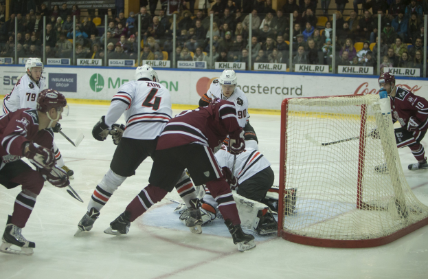 Eine Gruppe von Menschen, die auf einem Eisplatz Hockey spielt, trägt Sportkleidung, Helme und hält Hockeystöcke, mit einem Torpfosten auf der rechten Seite, umgeben von Brettern, Pfosten und Zuschauern in den Tribünen.