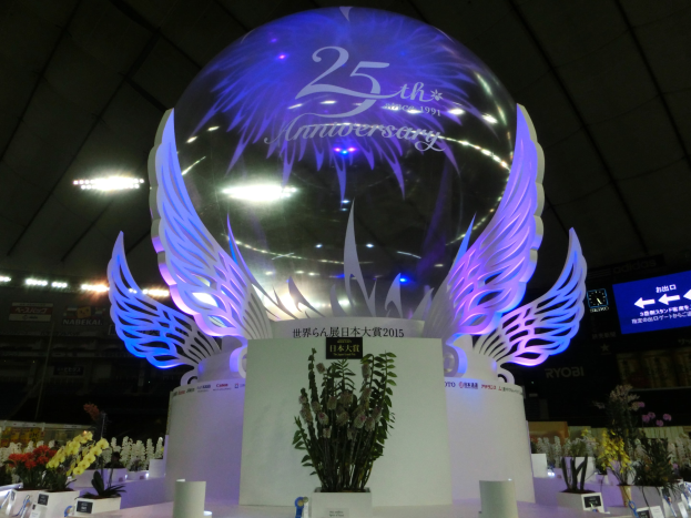 A large crystal ball with wings on top, surrounded by flower bouquets, name boards, and other objects on a festive floor, with boards, lights, and a screen in the background.