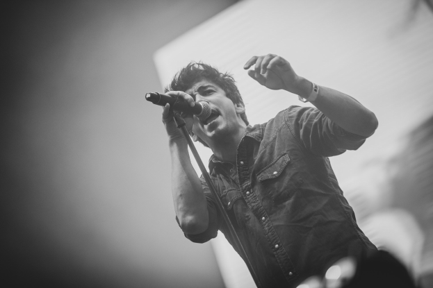 Schwarz-weißes Foto eines Mannes, der in ein Mikrofon singt, auf einer Bühne mit einem unscharfen Gesicht im Hintergrund.