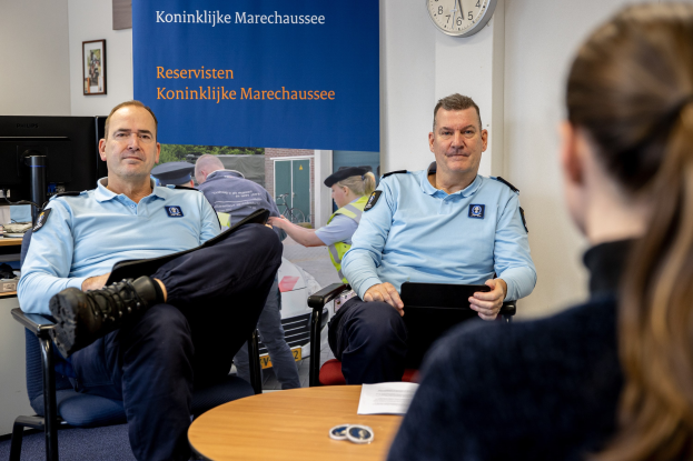 Zwei Polizeibeamte sitzen an einem Tisch mit einer Frau, Papieren und Gegenständen auf dem Tisch, einer Wandtafel und Uhr im Hintergrund, einem zweiten Tisch mit Monitor, Tastatur, Maus und Gegenständen links daneben und Fotorahmen an der Wand.