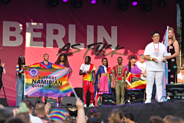 Eine Gruppe von Menschen auf einer Bühne mit einem Banner, auf dem 'Berlin as Fuck' steht, mit Lautsprechern, Mikrofonständen und einer feiernden Menge unten.