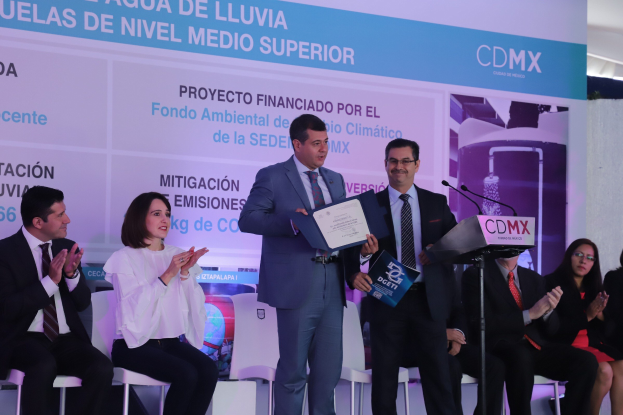 Eine Gruppe von Menschen, die auf Stühlen sitzen und auf ein Podium schauen, wo zwei Männer Zertifikate halten, mit einem Mikrofon auf dem Podium und einer Werbung im Hintergrund.