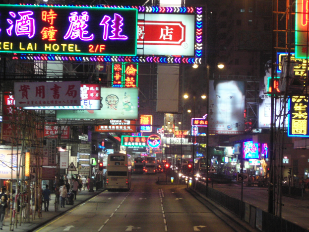 Eine nächtliche Stadtstraße mit fahrenden Fahrzeugen, Fußgängern auf dem Gehweg, beleuchteten Straßenlaternen, Gebäuden, Pfählen und verschiedenen Schildern und Plakaten.