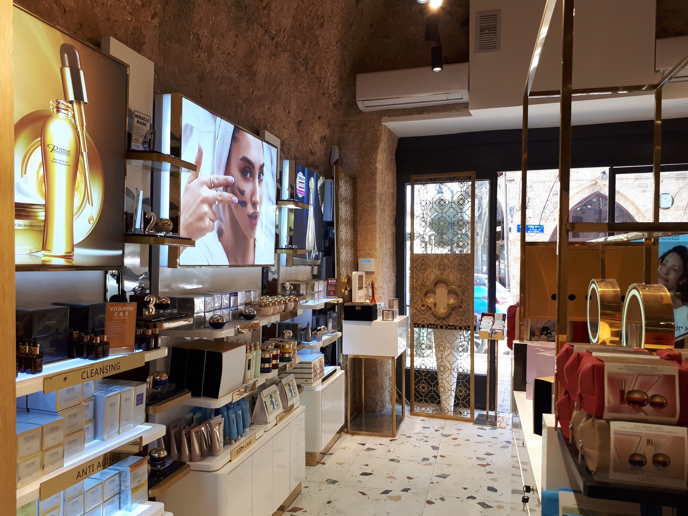 Innenansicht eines Kosmetikladens mit verschiedenen Produkten auf Regalen, Wandbildschirmen, Deckenleuchten, einer Klimaanlage und Glastüren, die eine Person, ein Auto und ein Gebäude draußen zeigen.