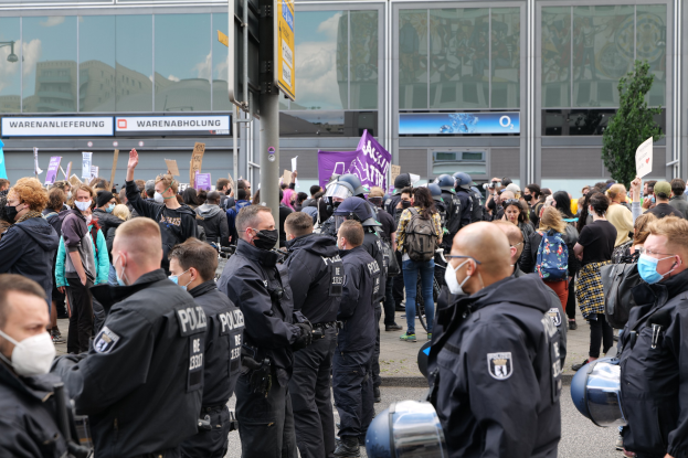 Eine große Gruppe von Menschen steht vor einem Gebäude und hält Schilder hoch, trägt Helme und hat eine Schilderstange im Vordergrund und einen Baum im Hintergrund, was auf eine Demonstration hindeutet.