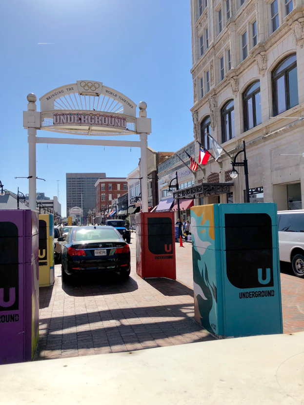 Stadtstraße mit parkenden Autos, einem Torbogen, Gebäuden, Laternen, Fahnenmasten, einem Müllcontainer und Himmel im Hintergrund.