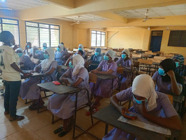Ein Klassenzimmer in Nigeria mit Schülerinnen und Schülern, die Gesichtsmasken tragen und auf Bänken sitzen oder stehen, umgeben von Büchern und Gegenständen auf Tischen, mit Fenstern, einer Tür, Lampen und Ventilatoren im Hintergrund.