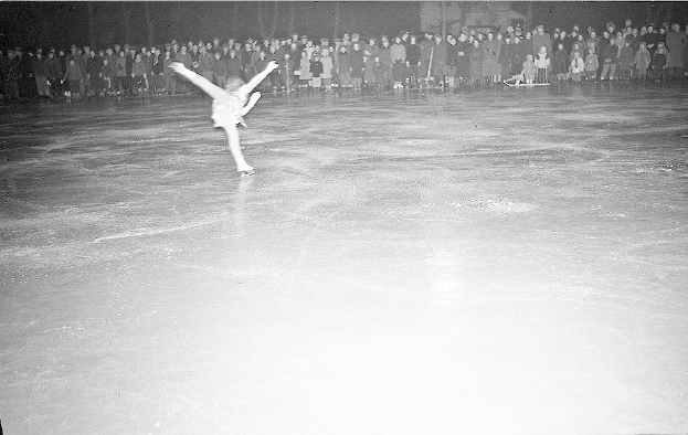 Ein Schwarz-Weiß-Foto eines Eiskunstläufers, der elegant über das Eis gleitet, während im Hintergrund eine Gruppe von Menschen steht.