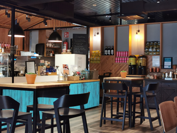 Inneneinrichtung eines Restaurants mit hölzernen Tischen, Stühlen, einem Blumentopf mit einer Pflanze auf einem Tisch, einer Schiefertafel mit Menü, einem Kühlschrank, einem Feuerlöscher, Regalen mit Flaschen, Schäubern mit Gegenständen, einem gerahmten Foto an der Wand und Deckenlampen.