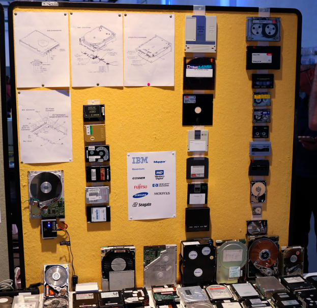 Ein Tisch mit verschiedenen Festplatten darauf, daneben steht eine Person und hält ein Objekt in der Hand, im Hintergrund ist eine Tafel mit Papieren zu sehen.
